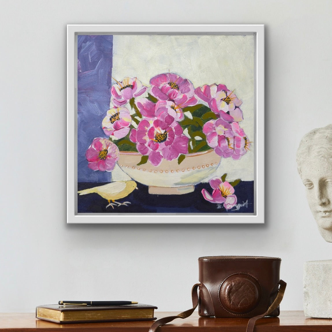 Framed artwork of pink flowers in a bowl on a wall above a desk with a camera and books for art collectors, home decor.