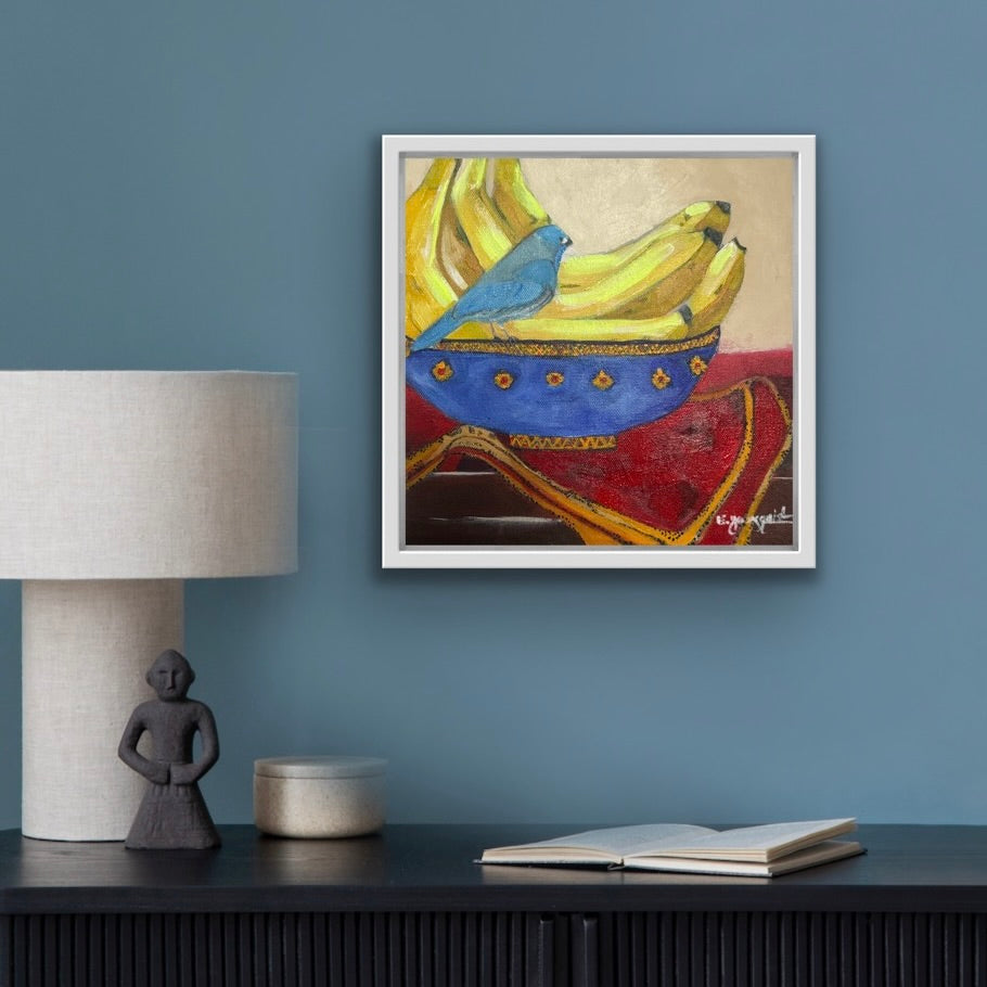 Framed artwork of a blue bird, bananas, displayed on a wall above a table with decor items.