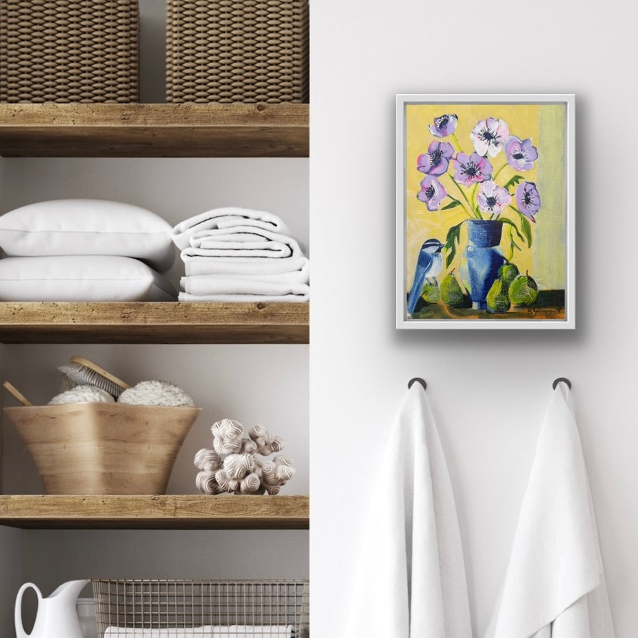 Framed artwork of a blue vase with purple flowers on a white wall above a shelf with towels and a basket.