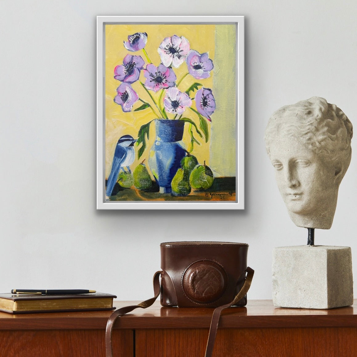 Framed original still life painting of bird and flowers on a wall above a wooden surface with a camera and sculpture.