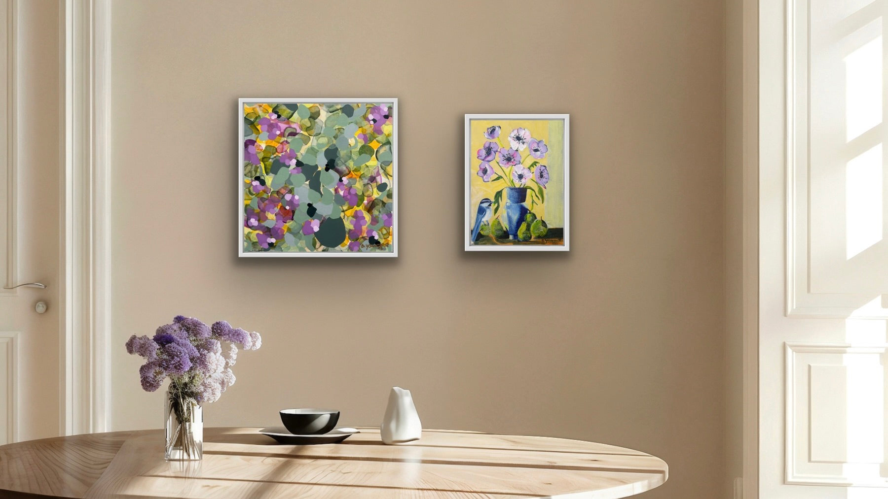 Two floral paintings, a still life and an abstract on a beige wall above a wooden table with decorative items.